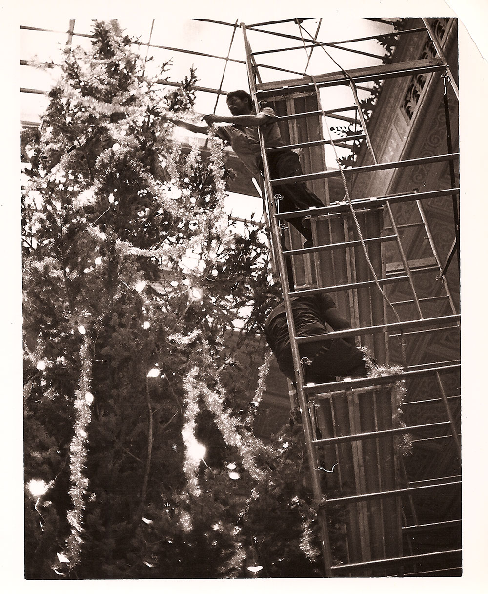 (FNB.2010.2.05) - Two Men Hang Garland on Christmas Tree in the Great Banking Hall, First National Center, c. 1974